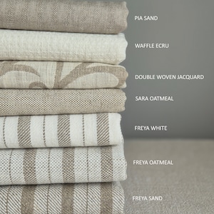 Op de afbeelding: Een stapel van acht verschillende linnen stoffen in verschillende tinten beige en wit. De stoffen zijn gelabeld met hun namen: Pia Sand, Waffle Ecru, Double Woven Jacquard, Sara Oatmeal, Freya White, Freya Oatmeal en Freya Sand.