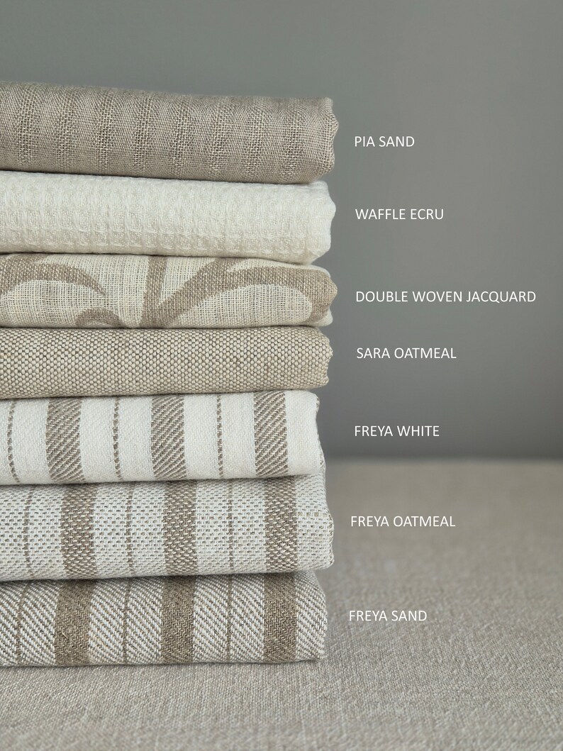 Op de afbeelding: Een stapel van acht verschillende linnen stoffen in verschillende tinten beige en wit. De stoffen zijn gelabeld met hun namen: Pia Sand, Waffle Ecru, Double Woven Jacquard, Sara Oatmeal, Freya White, Freya Oatmeal en Freya Sand.