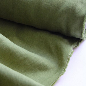 May include: A roll of olive green fabric with a visible woven texture. The material is partially unrolled, revealing soft folds and a raw, frayed edge along the side. The fabric displays a consistent, earthy green tone throughout the entire piece.