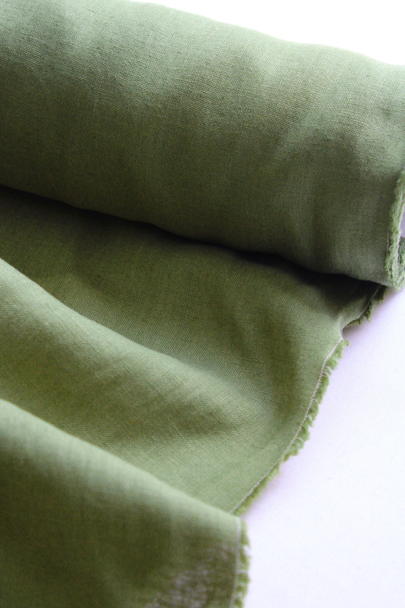May include: A roll of olive green fabric with a visible woven texture. The material is partially unrolled, revealing soft folds and a raw, frayed edge along the side. The fabric displays a consistent, earthy green tone throughout the entire piece.