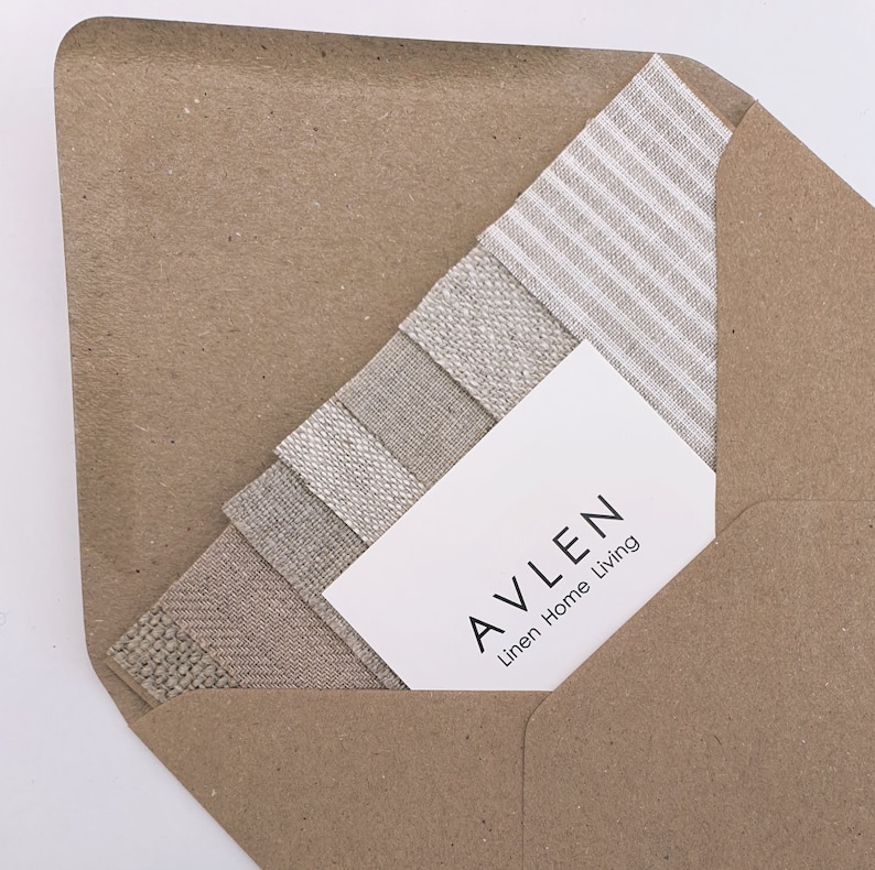 Op de afbeelding: Een witte visitekaartje met de tekst "AVLEN Linen Home Living" in een bruine papieren envelop. De envelop is gedeeltelijk gevuld met stofstalen van verschillende linnen stoffen in verschillende tinten beige en wit.