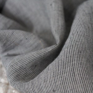 May include: A piece of fabric featuring a fine pinstripe pattern in shades of charcoal gray and off-white. The material has a visible woven texture with subtle variations in the thread, displayed in soft, draped folds that create depth and shadow.