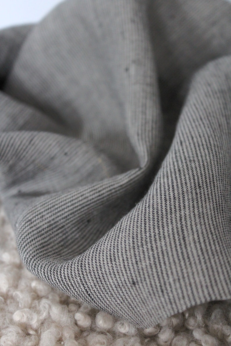 May include: A piece of fabric featuring a fine pinstripe pattern in shades of charcoal gray and off-white. The material has a visible woven texture with subtle variations in the thread, displayed in soft, draped folds that create depth and shadow.