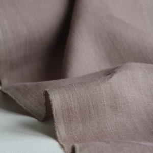May include: A length of dusty rose fabric with a visible woven texture and a raw, unfinished edge. The material is draped in soft folds, highlighting the matte finish and the muted, earthy pink tone of the textile surface throughout the piece.