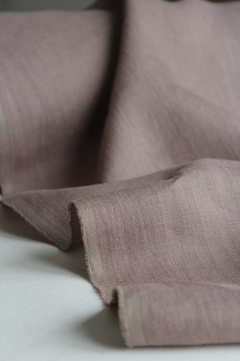 May include: A length of dusty rose fabric with a visible woven texture and a raw, unfinished edge. The material is draped in soft folds, highlighting the matte finish and the muted, earthy pink tone of the textile surface throughout the piece.