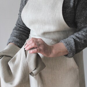 May include: A natural linen apron with adjustable straps and a large pocket. The apron is a light beige colour and is made of a soft, natural fabric.