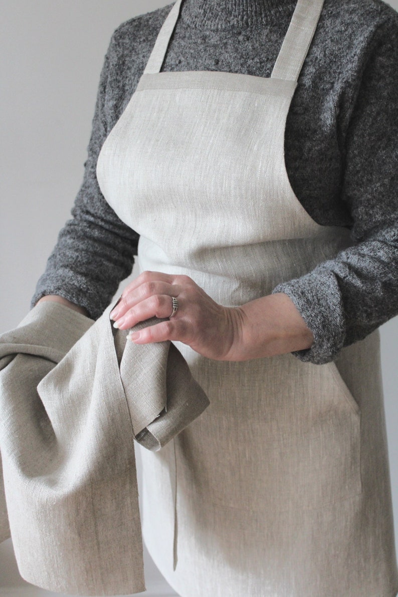 May include: A natural linen apron with adjustable straps and a large pocket. The apron is a light beige colour and is made of a soft, natural fabric.