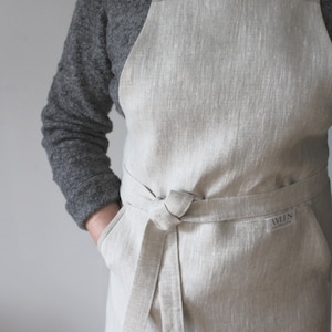 May include: A beige linen apron with adjustable straps and a large pocket. The apron has a tie waist and a small label that says "AWLN".