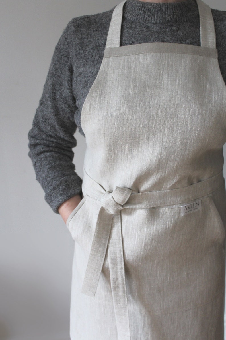 May include: A beige linen apron with adjustable straps and a large pocket. The apron has a tie waist and a small label that says "AWLN".