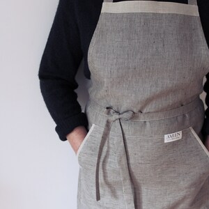 May include: A grey and white striped linen apron with a white trim and a large pocket on each side. The apron has a long tie that can be tied at the waist. The apron has a small white label with the text "AVLN" on it.