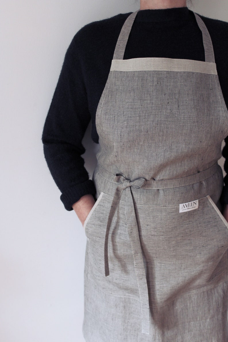 May include: A grey and white striped linen apron with a white trim and a large pocket on each side. The apron has a long tie that can be tied at the waist. The apron has a small white label with the text "AVLN" on it.