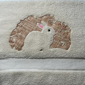 May include: Cream-colored bath towel with an embroidered design of a white rabbit with pink ears and a brown leafy background. The towel has a textured surface and a white band near the bottom.