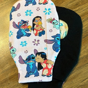 May include: A pair of black oven mitts with a light pink patterned overlay. The overlay features blue cartoon characters, flowers, and a girl in a red dress. The mitts are likely for kitchen use.