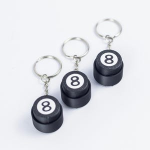 Könnte beinhalten: Drei Schlüsselanhänger mit schwarz-weißem 8-Ball-Design. Jeder Schlüsselanhänger hat eine schwarze, zylindrische Basis mit einem weißen Kreis oben, der die Zahl 8 in Schwarz anzeigt. Die Schlüsselanhänger haben silberne Schlüsselringe.