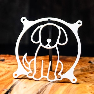 May include: A white, outline-style dog ornament, sitting within a circular frame. The dog has large, floppy ears and a curled tail. The frame features four small holes, possibly for hanging or attaching to something.
