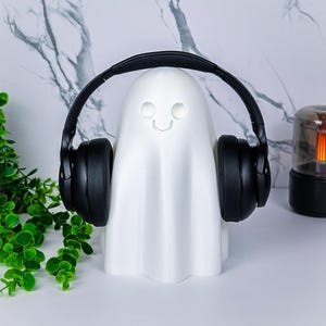 May include: A white ghost-shaped headphone stand with a smiling face, wearing black headphones. The stand is on a white surface with a marble-patterned background and a small green plant.