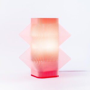 A modern, geometric table lamp. The lamp features a layered, pleated design with a rectangular shape. The base is a solid red, and the lamp emits a warm, diffused light.