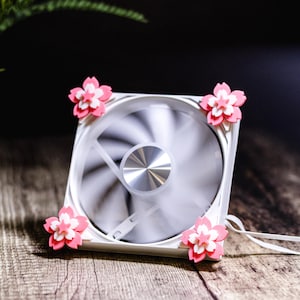 Peut inclure: Un ventilateur d'ordinateur blanc avec des pales en rotation, orné de quatre décorations florales roses et blanches à chaque coin. Le ventilateur est posé sur une surface en bois, avec un câble d'alimentation.