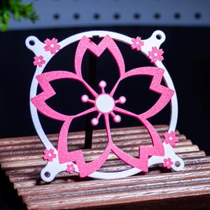May include: A decorative, laser-cut cherry blossom design in pink and white. The flower is centered within a white circular frame, with smaller pink blossoms around the edges. The design is displayed on a wooden stand.