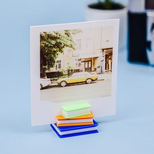 May include: A square white photo print featuring a yellow car parked on a city street, held upright by a stack of four miniature plastic books in lime green, orange, yellow, and blue, serving as a decorative desk stand for displaying photographs.