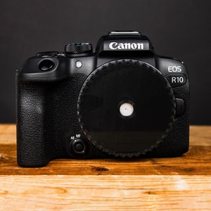 May include: A black Canon EOS R10 camera with a lens cap on the front. The camera has a black body with white text and a black lens.