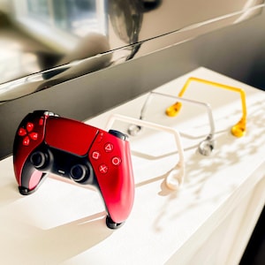 Puede incluir: Un mando de videojuegos rojo y negro con un soporte a juego en primer plano. Tres soportes en blanco, plateado y dorado están detrás del mando. El mando tiene un centro negro y laterales rojos con la disposición estándar de botones.