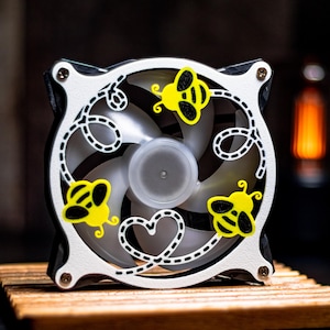 May include: A decorative computer fan with a white frame and black accents. The fan blades are visible behind a white design featuring yellow bees and a heart shape. The fan sits on a wooden surface.