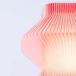 A close-up of a modern, abstract sculpture in red. The lamp has a ribbed, layered design, with vertical lines creating a textured surface. The sculpture's form is geometric, with a curved, organic shape.