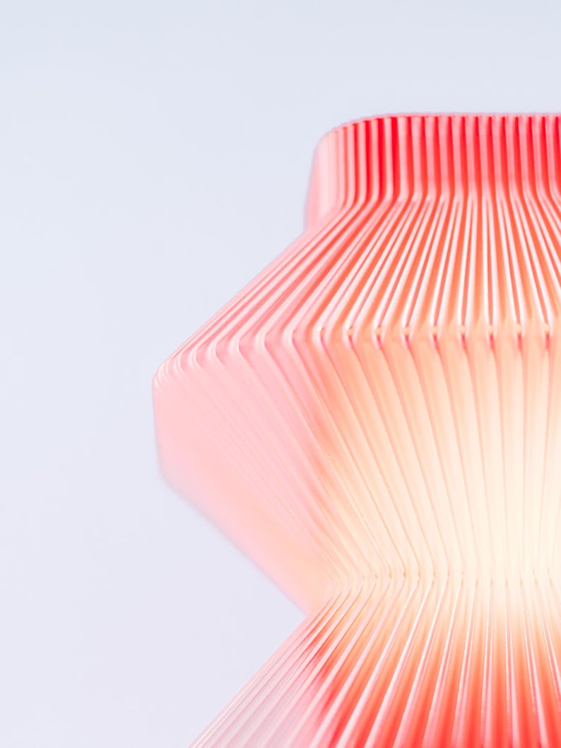 A close-up of a modern, abstract sculpture in red. The lamp has a ribbed, layered design, with vertical lines creating a textured surface. The sculpture's form is geometric, with a curved, organic shape.