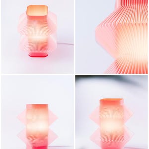 A modern, geometric table lamp in red. The lamp features a layered, pleated design with a square top and base. The lamp is lit, emitting a warm glow, and is set against a white background.