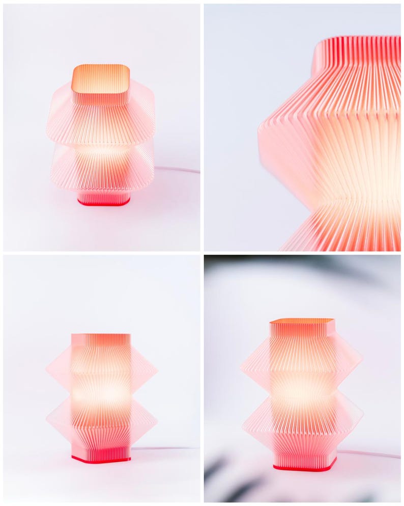 A modern, geometric table lamp in red. The lamp features a layered, pleated design with a square top and base. The lamp is lit, emitting a warm glow, and is set against a white background.