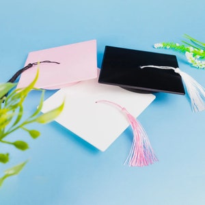 May include: Three graduation caps in pink, black and white with matching tassels. The caps are arranged on a light blue background.
