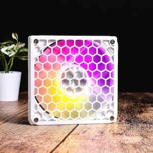 Pode incluir: Uma ventoinha de arrefecimento para computador de formato quadrado com moldura branca e grelha hexagonal em favo de mel branca. As pás são iluminadas com um gradiente vibrante de luzes LED amarelas, rosas e roxas, criando um brilho circular.