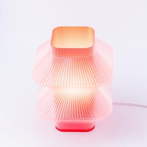 A red, tiered table lamp with a ribbed design. The lamp has a square top and a square base, with a warm glow emanating from within. The lamp is made of a translucent plant based plastic and has a modern aesthetic.