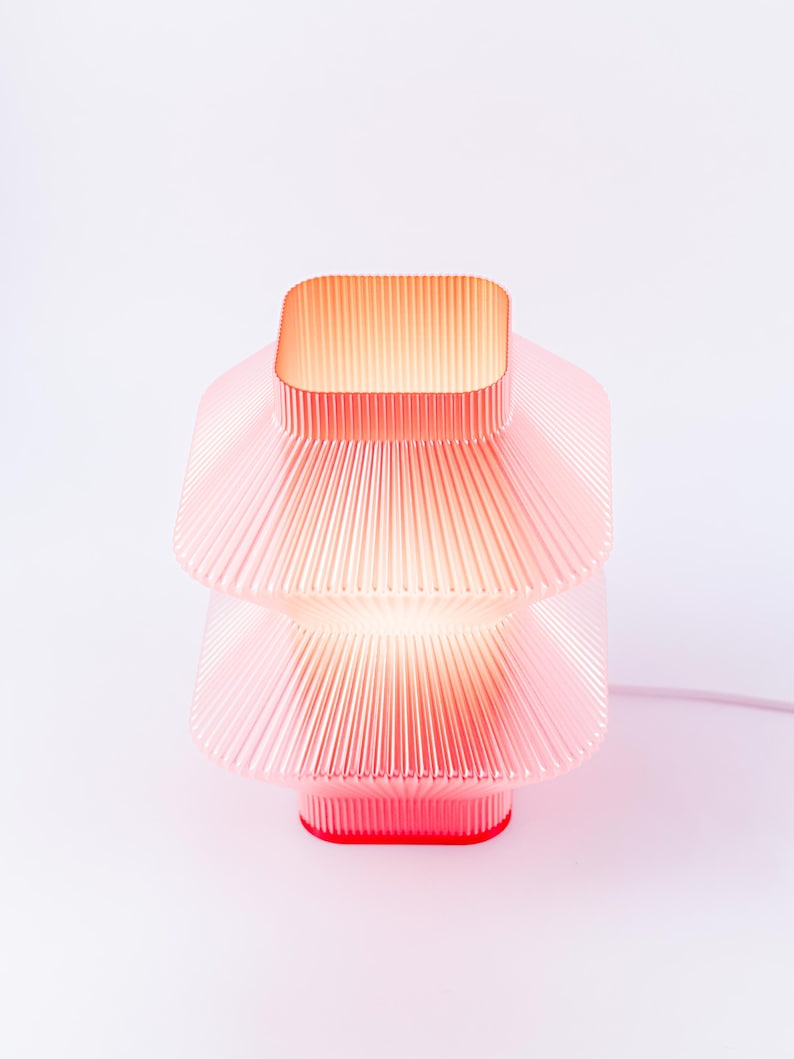 A red, tiered table lamp with a ribbed design. The lamp has a square top and a square base, with a warm glow emanating from within. The lamp is made of a translucent plant based plastic and has a modern aesthetic.