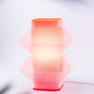 A red, geometric table lamp with a pleated design. The lamp has a square base and top, with a stacked, diamond-like shape in the middle. The lamp is lit from within, creating a warm glow. The cord is white.