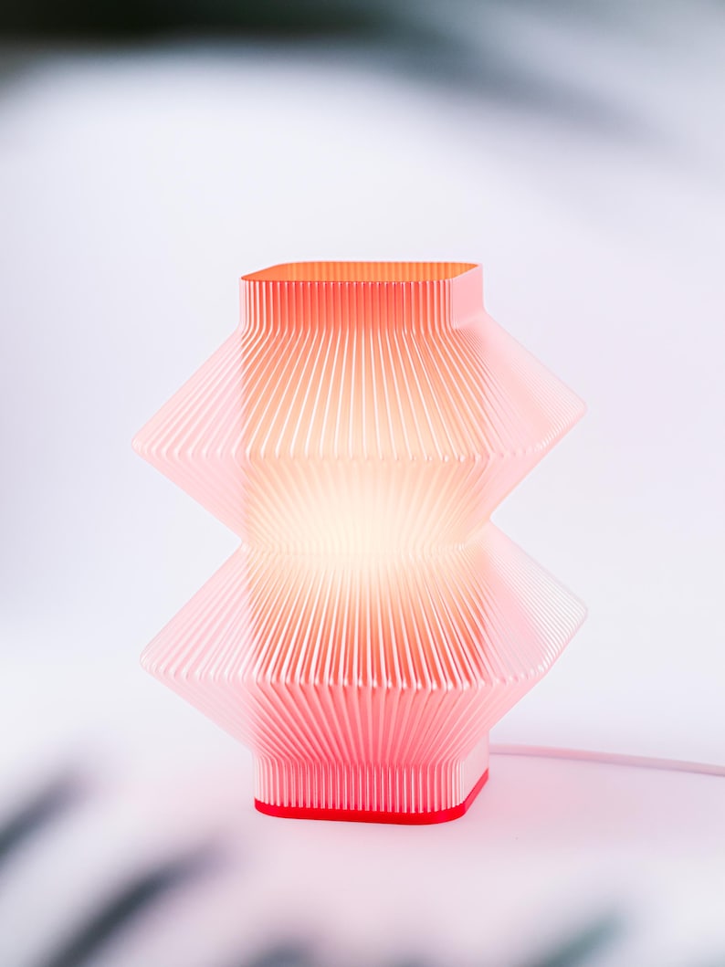 A red, geometric table lamp with a pleated design. The lamp has a square base and top, with a stacked, diamond-like shape in the middle. The lamp is lit from within, creating a warm glow. The cord is white.