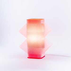 A red geometric table lamp with a pleated design. The lamp has a square top and base, with a diamond-shaped center. The lamp is lit, emitting a warm glow. The lamp is connected to a white power cord.