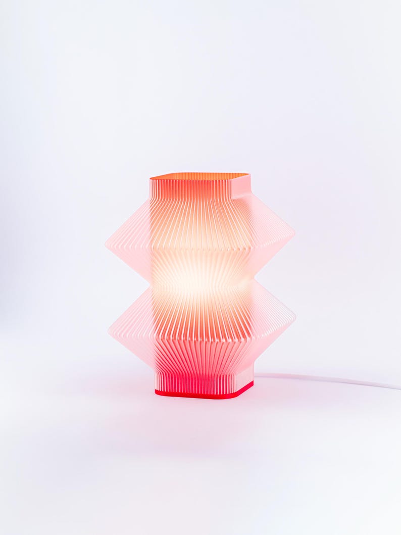 A red geometric table lamp with a pleated design. The lamp has a square top and base, with a diamond-shaped center. The lamp is lit, emitting a warm glow. The lamp is connected to a white power cord.