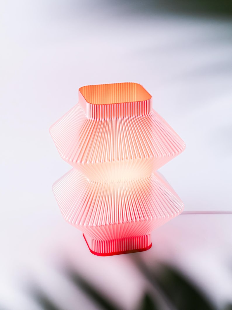 A red, geometric table lamp with a pleated design. The lamp has rectangular shape with a red base and a warm, red glow emanating from within.