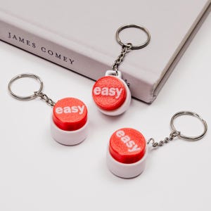 May include: Three white keyrings with red buttons that say "easy" in white letters. The keyrings are on a white surface.