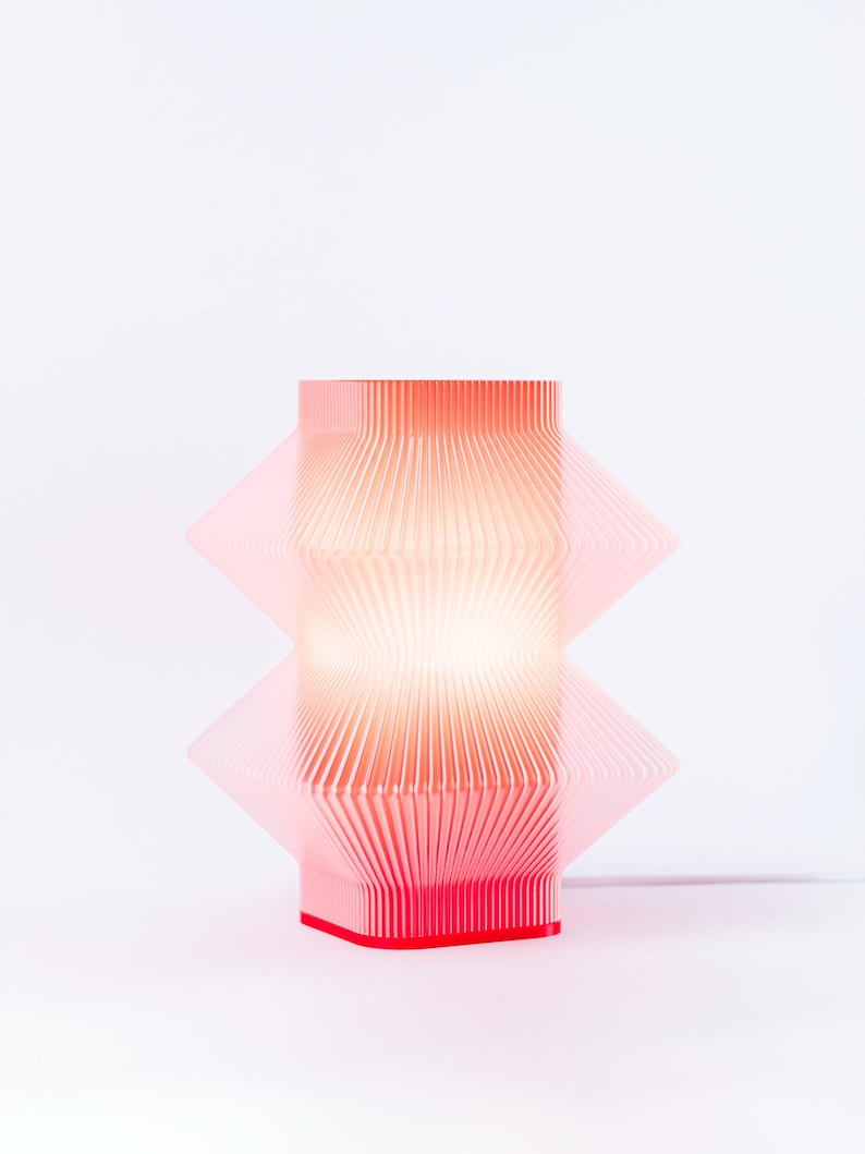 A modern, geometric table lamp. The lamp features a layered, pleated design with a rectangular shape. The base is a solid red, and the lamp emits a warm, diffused light.