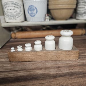 May include: A collection of vintage white ceramic weights displayed in a wooden tray. The weights, with rounded tops, are arranged by size. The wooden tray has individual slots for each weight. The weights are likely measured in grams.