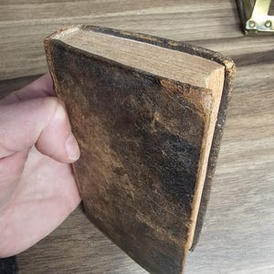 K&ouml;nnte beinhalten: Ein antikes Buch mit einem abgenutzten, dunkelbraunen Ledereinband. Die Kanten der Seiten sind sichtbar und zeigen eine hellbraune Farbe. Das Buch wird in einer Hand gehalten, was auf seine Gr&ouml;&szlig;e und sein Alter hindeutet.