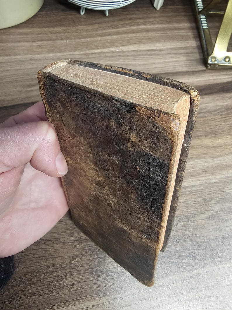 K&ouml;nnte beinhalten: Ein antikes Buch mit einem abgenutzten, dunkelbraunen Ledereinband. Die Kanten der Seiten sind sichtbar und zeigen eine hellbraune Farbe. Das Buch wird in einer Hand gehalten, was auf seine Gr&ouml;&szlig;e und sein Alter hindeutet.
