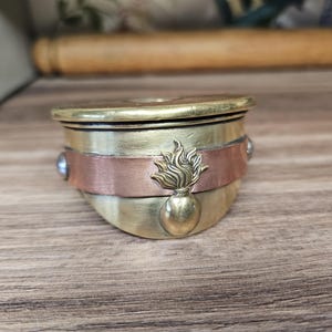 Op de afbeelding: Een kleine, ronde, messingkleurige container met een koperen band en een decoratief vlammenembleem. De container heeft een deksel en is geaccentueerd met kleine, ronde, blauwgrijze stenen. Het item staat op een houten oppervlak.