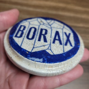 May include: A white ceramic lid with a blue border and the word "BORAX" printed in blue letters. The lid has a cracked glaze.