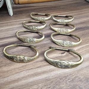 Puede incluir: Ocho servilleteros antiguos de latón con forma ovalada y un diseño central decorativo. Los anillos están dispuestos sobre una superficie de madera, mostrando su pátina envejecida y sus intrincados detalles. Un artículo de decoración vintage.