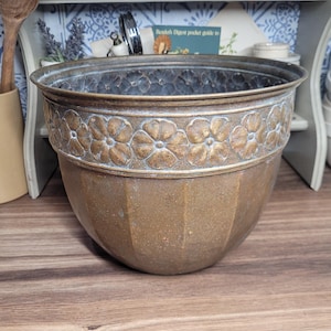 May include: A large, round, brass planter with a decorative floral design around the top rim. The planter has a slightly distressed, aged look.