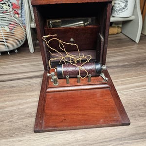Puede incluir: Una caja de madera antigua con tapa con bisagras, que revela compartimentos internos y un componente cilíndrico envuelto en cuero marrón. La caja tiene un asa y está abierta, mostrando cables y accesorios metálicos. La madera tiene un acabado oscuro y rico.